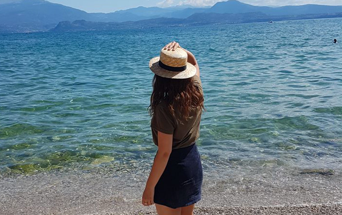 a woman stands on the edge of the sea. She holds on to her hat.