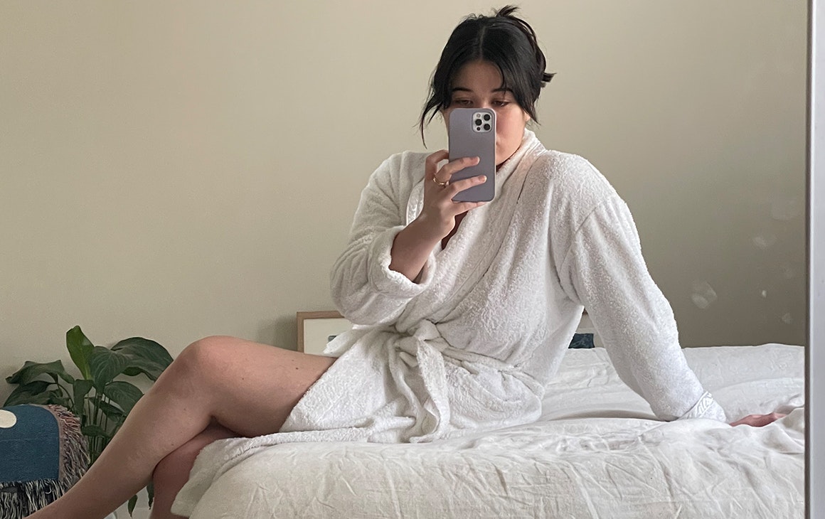 a woman sits on the best in a bathrobe, taking a selfie.