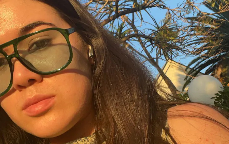 a close up selfie of a woman wearing sunglasses, a blue sky behind her.