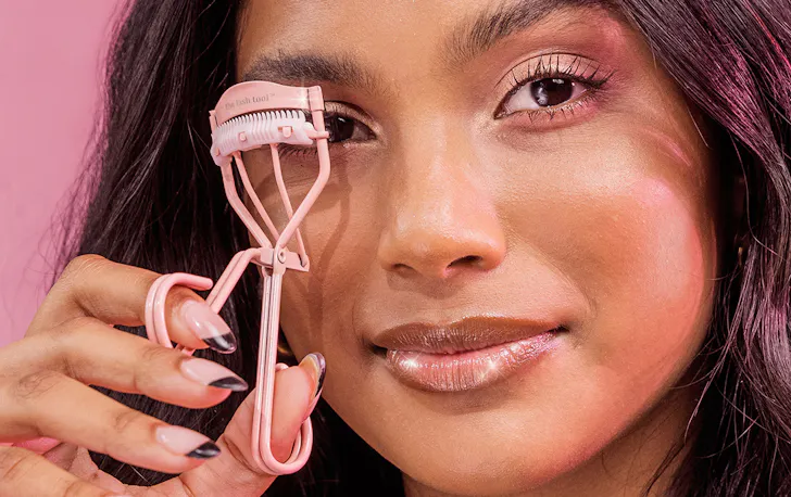 a woman holds up a pink eyelash curler up to her eye.