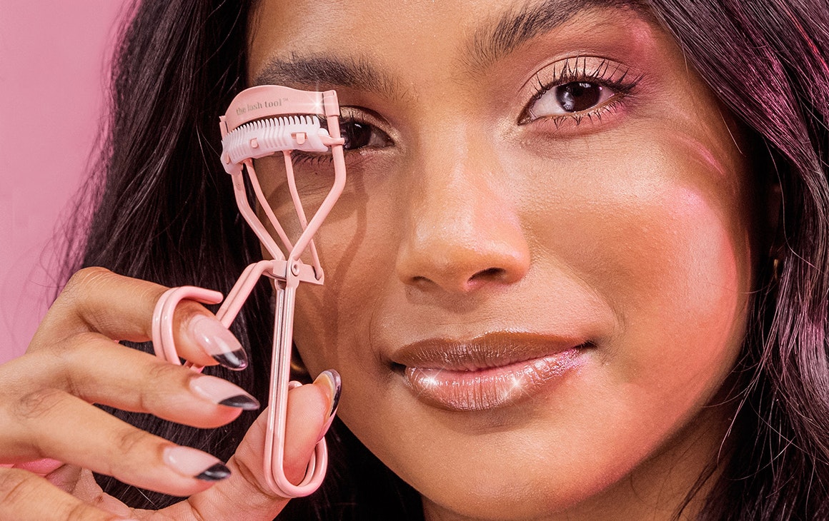 a woman holds up a pink eyelash curler up to her eye.