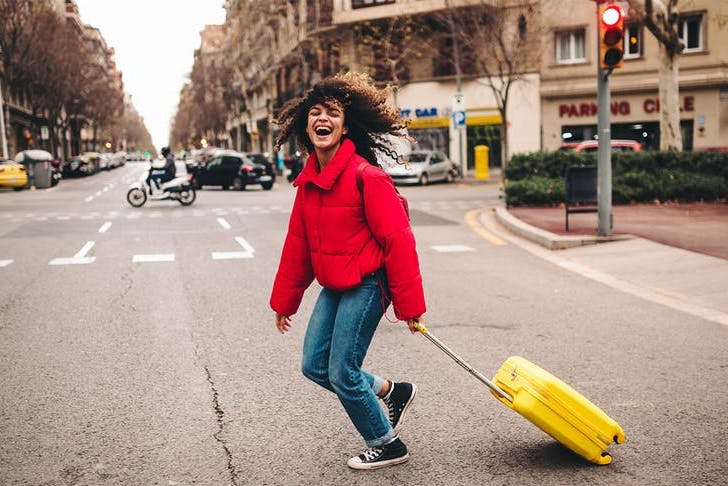 a woman in a red jacket laughs as she pulls a yellow suitcase across the street