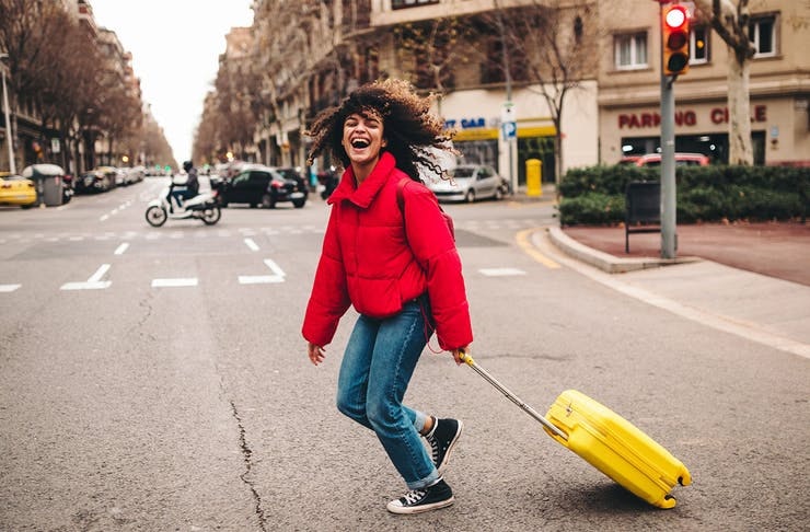 a woman in a red jacket laughs as she pulls a yellow suitcase across the street