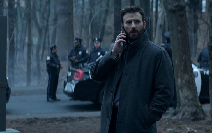 A man talks on his phone in the middle of a park on a gloomy day. Behind him is a cluster of police.