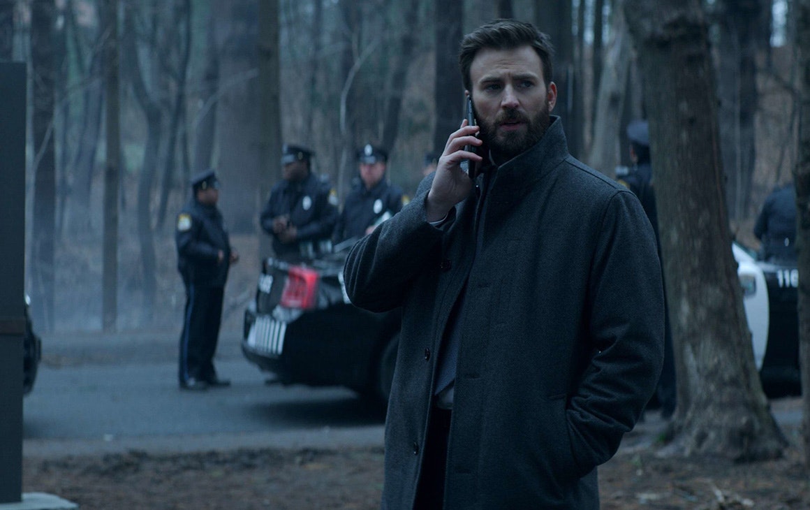 A man talks on his phone in the middle of a park on a gloomy day. Behind him is a cluster of police.