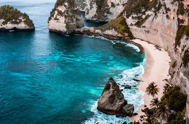 a beach in bali shows off white sand, blue sea and a rugged rocky coastline.