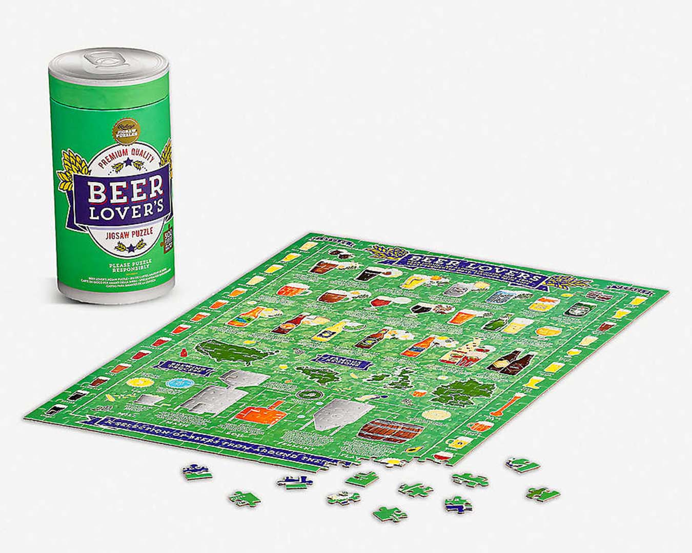 Green, partially made puzzle featuring pictures of beer, sits on a white table. In the background is a puzzle box in the shape of a beer tin.