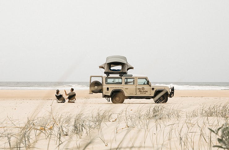 a four wheel drive parked up on a beach, with two people sitting in camping chairs next to it