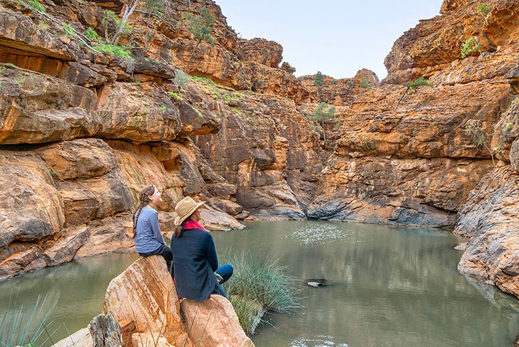 two people sitting on rock looking out onto a bush creek