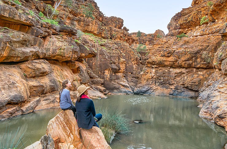 two people sitting on rock looking out onto a bush creek