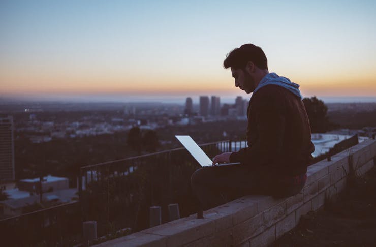man sitting on top of roof with skyline of city holding laptop