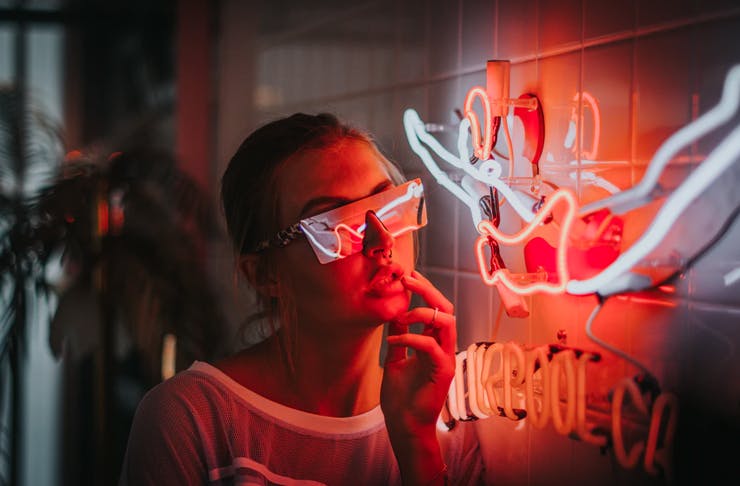 young woman wearing futuristic screen glasses leaning next to neon sign hanging on tiled wall