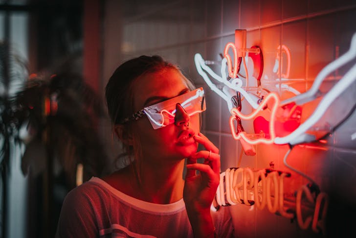 young woman wearing futuristic screen glasses leaning next to neon sign hanging on tiled wall