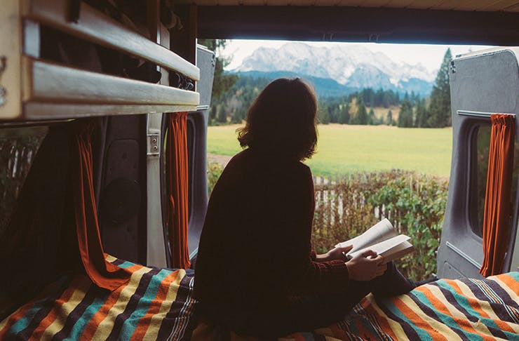 girl sitting inside travel van with book and van back doors open overlooking mountainous landscape