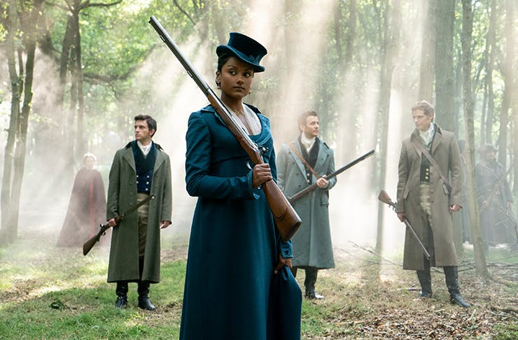 lady standing in the woods in period clothing, holding a gun, three men are standing behind her