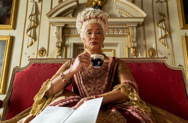 Queen Charlotte of Bridgerton holds a paper and sips a cup of tea.