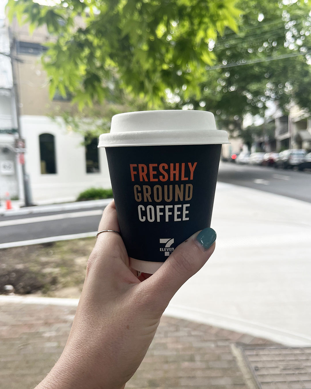 A hand holding a 7-Eleven coffee with trees and a footpath in the background