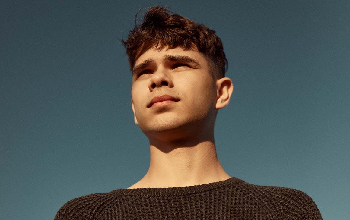 Singer Budjerah, wearing a dark jumper, stares off into the distance. A blue sky behind him.