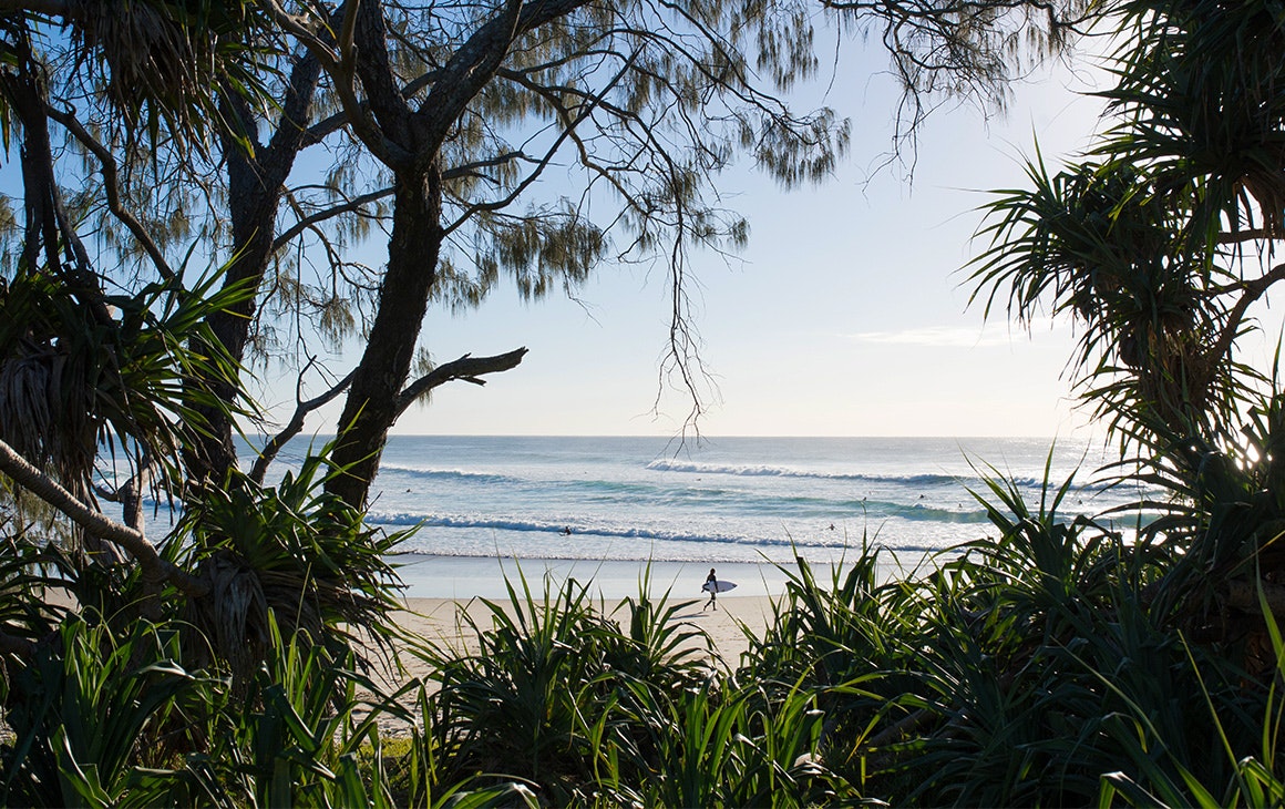 clusters of trees frame an image of a white sand beach. A person walks in the distance.