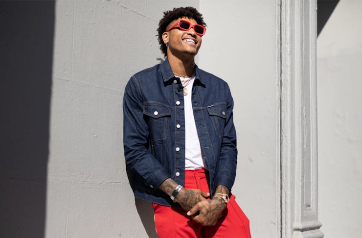 a man in red jeans and matching red sunglasses laughs while leaning against a wall.