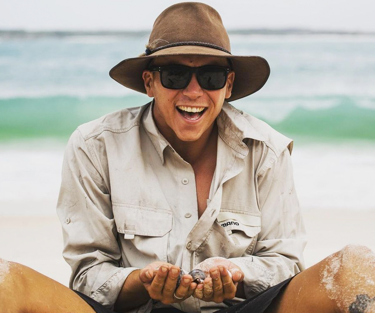 young man sitting on the beach wearing a wide brim hat and holding a crab laughing