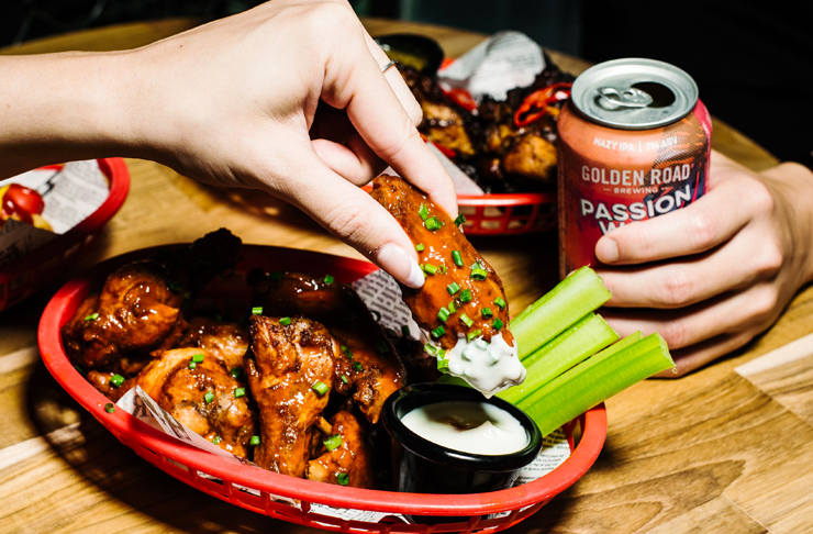 hand reaching into chicken wing basket and dipping into sauce with another hand holding a beer