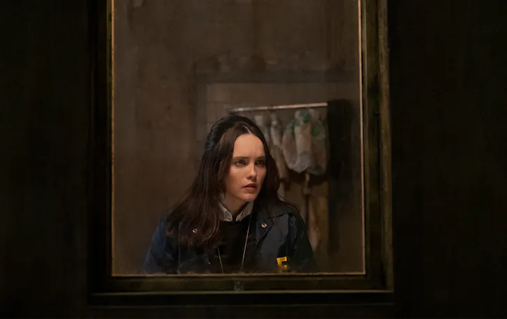 a young woman, peers through a dark window.