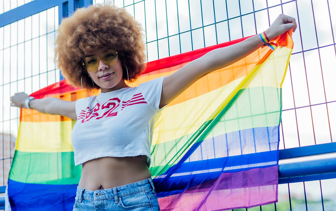 a person wearing a white crop top and jeans proudly holds up a rainbow flag