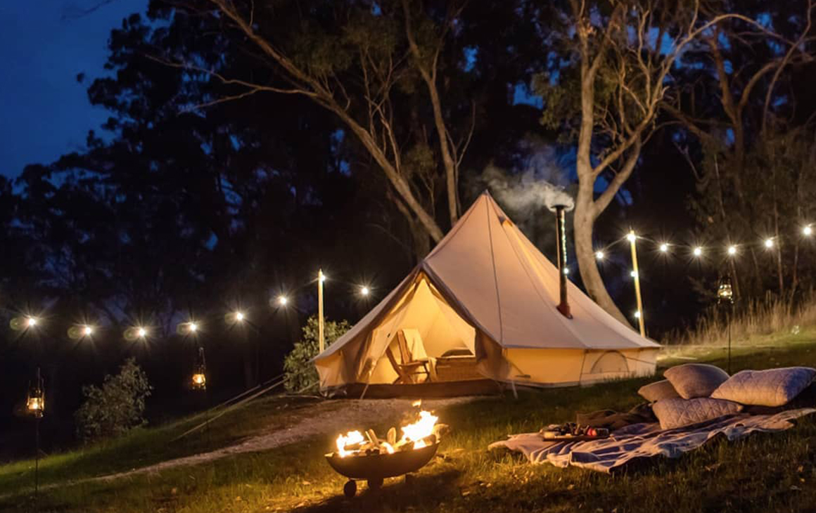 a string of fairy lights hang from a tent at night. In front of it is a crackling fire.