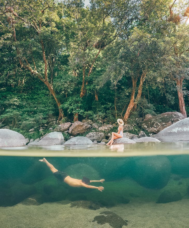 a man swims in the crystal clear water of Mossman Gorge. A woman lounges on a rock.