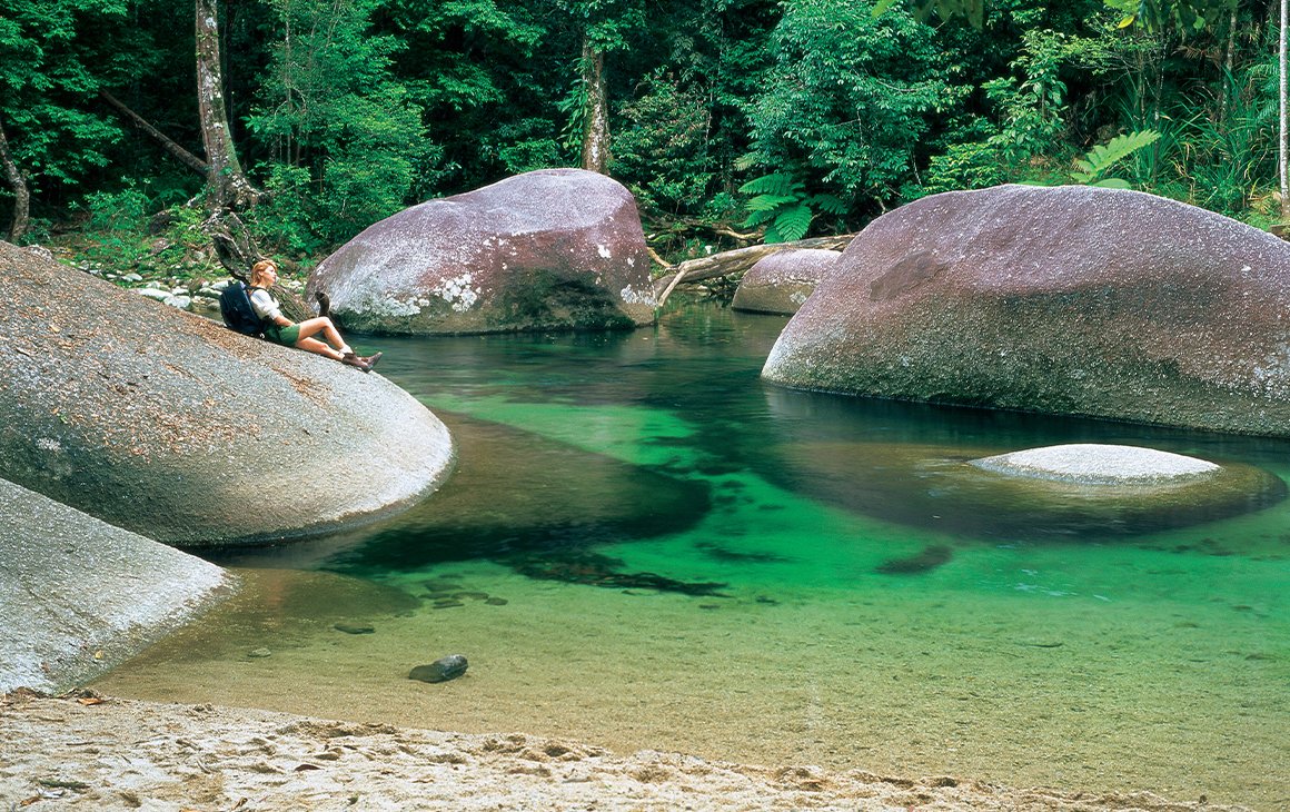 Girl resting on large smooth boulder which is slightly submerged in a crystal clear lake and surrounded by bush land