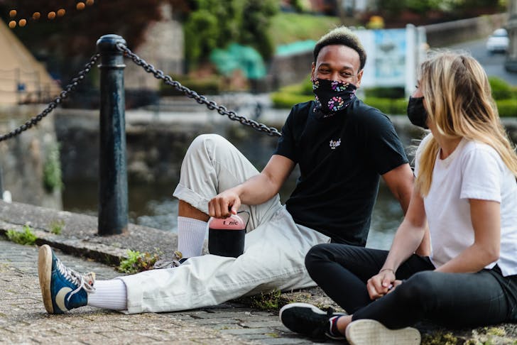 A guy and a girl sitting down on the ground wearing face masks. 
