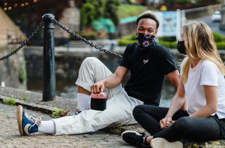 A guy and a girl sitting down on the ground wearing face masks. 