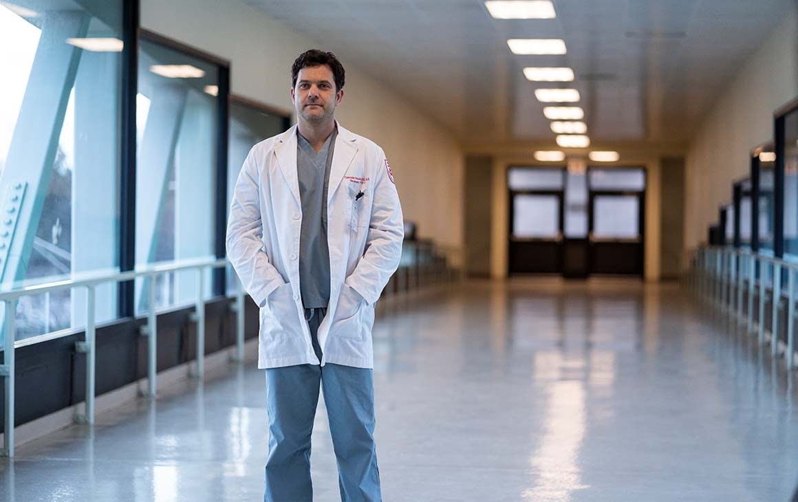 A doctor stands in the hallway of a hospital, his hands in the pocket of his jacket.