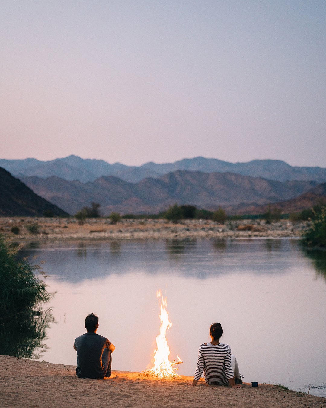 two people sitting on edge of river bank with small fire, looking out onto river at sunset