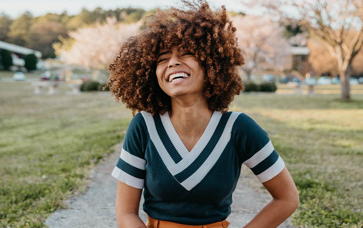 a young woman, sits in a park and throws her head back laughing