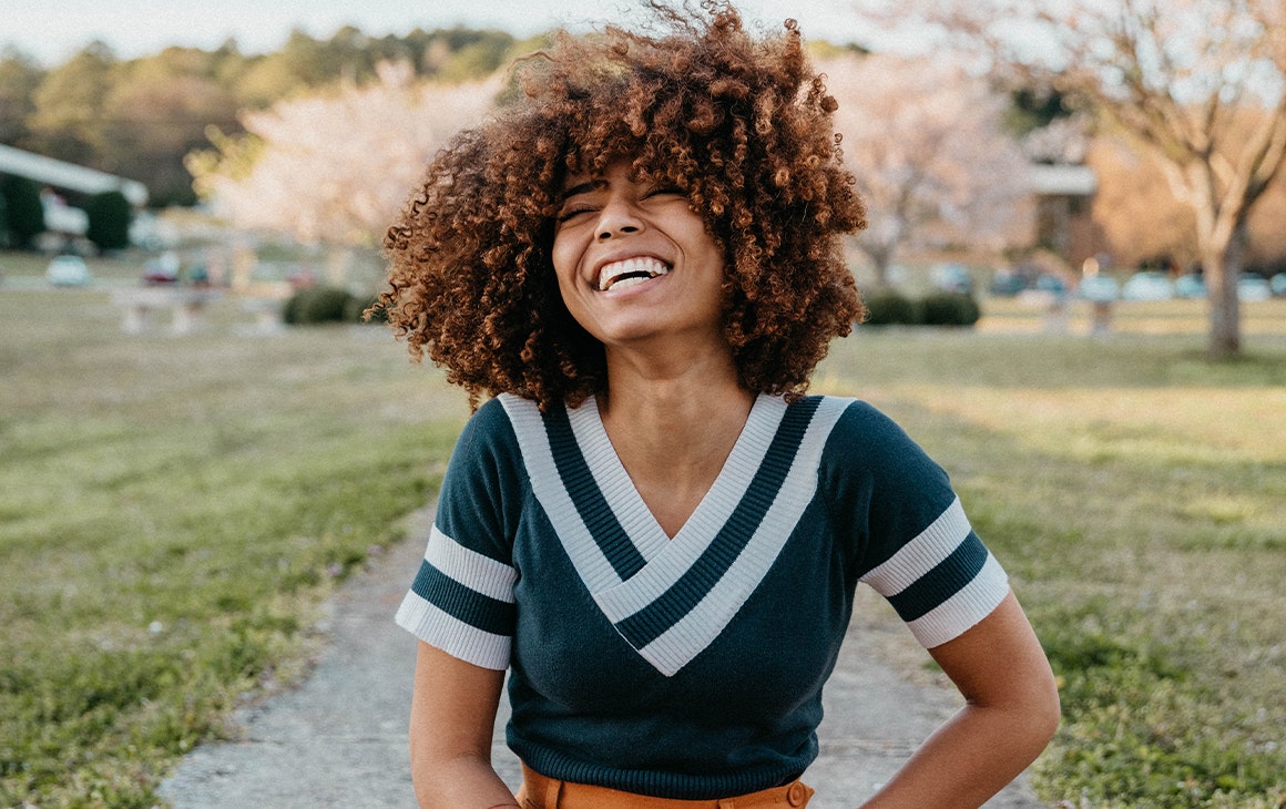 a young woman, sits in a park and throws her head back laughing