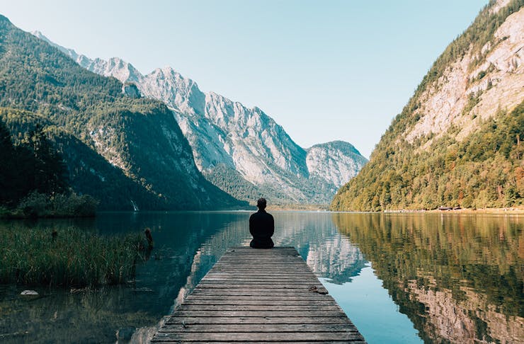 a person sits on the end of a dock by a lake and mountains.
