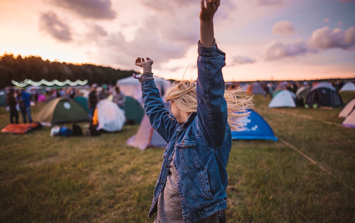 a young woman throws her arms up in the air. A cluster of tents are behind her.