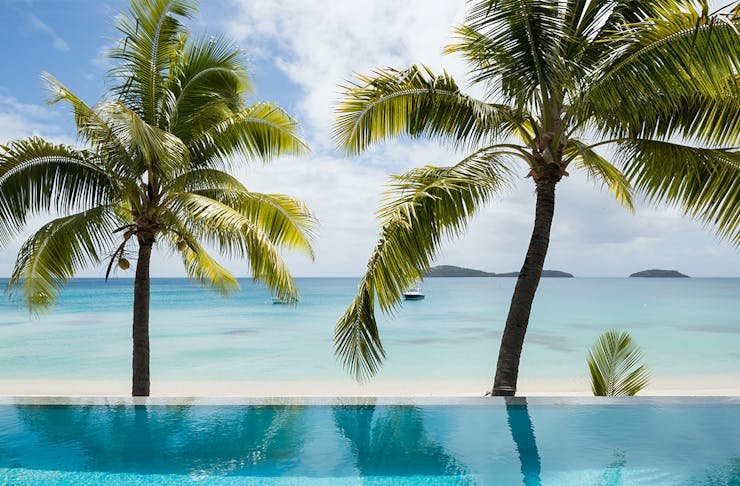 a pool sits next to the ocean, fringed by palm trees.