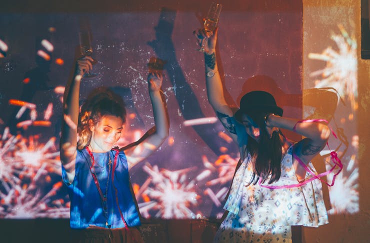 Two young women dance at a house party with a glass of wine in their hand. They're covered by a reflection of fireworks. 