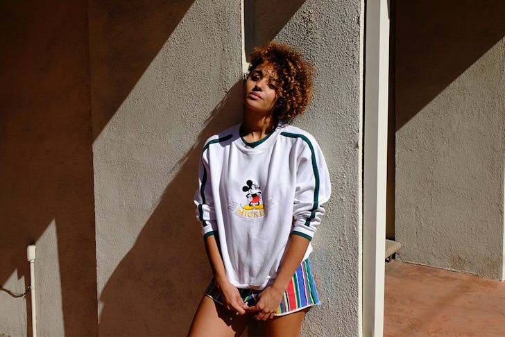 a woman in a Mickey Mouse jumper leans against a concrete wall, basking in the sun.