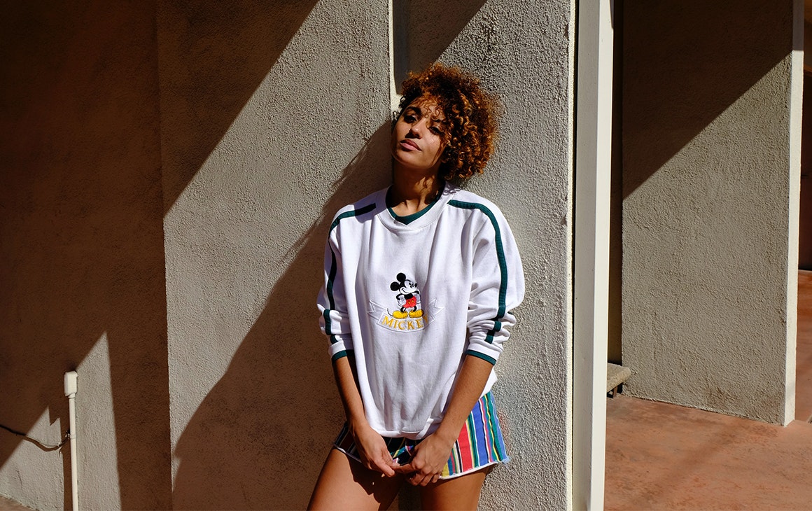 a woman in a Mickey Mouse jumper leans against a concrete wall, basking in the sun.