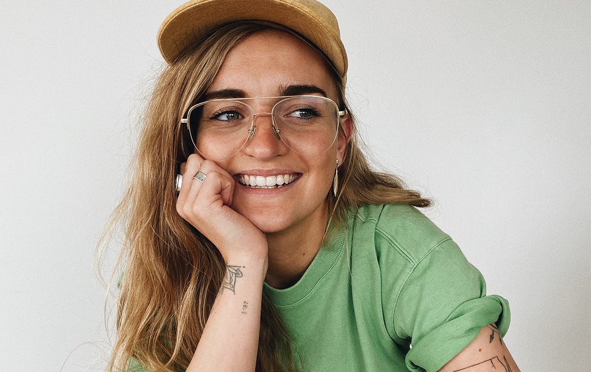 G Flip wearing a green tee, hat and glasses, rests her head on her hand, smiling.