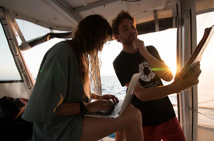 two people on a boat, one is on a laptop and the other is looking at a report