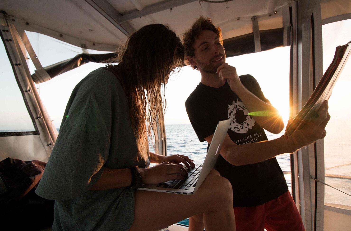 two people on a boat, one is on a laptop and the other is looking at a report