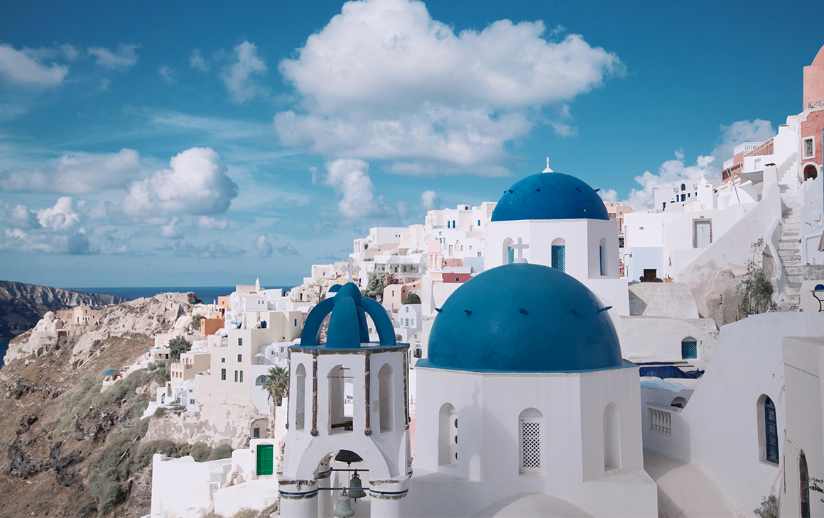 a cluster of white buildings with blue rooftops cover a cliffside in Greece.