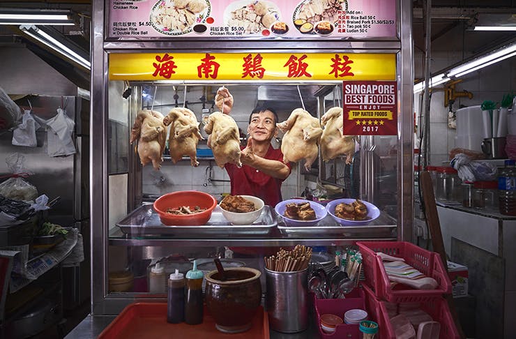 Man serving chicken at a hawker centre with chicken carcasses strung up above his head. 