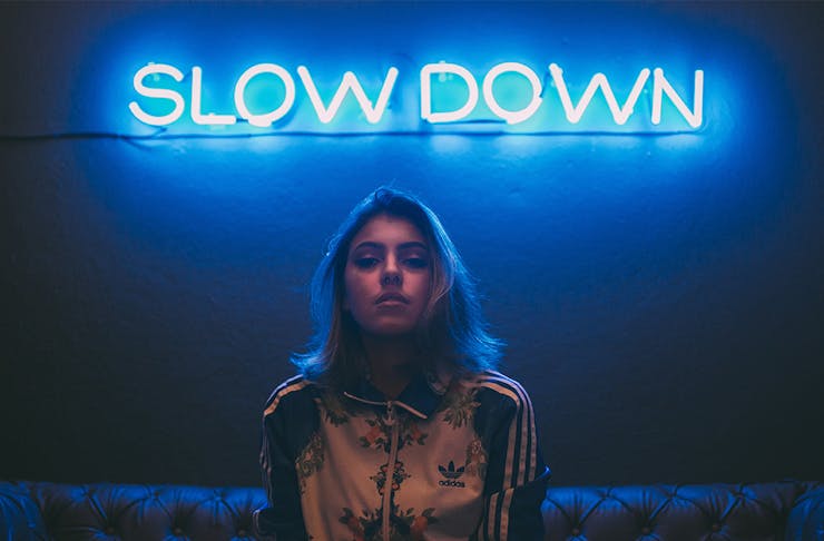 a young woman sits on a couch in front of a neon blue sign that says 'Slow Down'