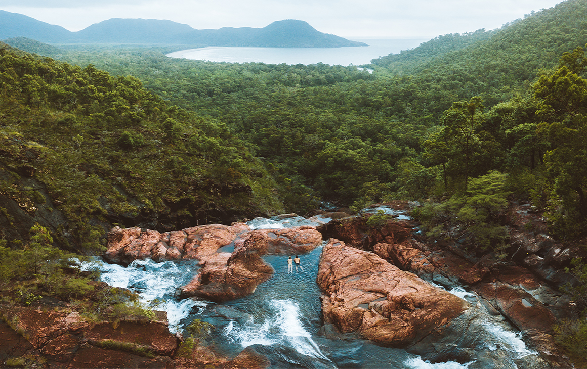 Aerial image of a two people floating in a rock pool looking over the rainforest